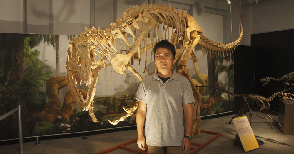 Yoshitsugu Kobayashi standing in front of the skeleton of an afrovenator dinosaur.