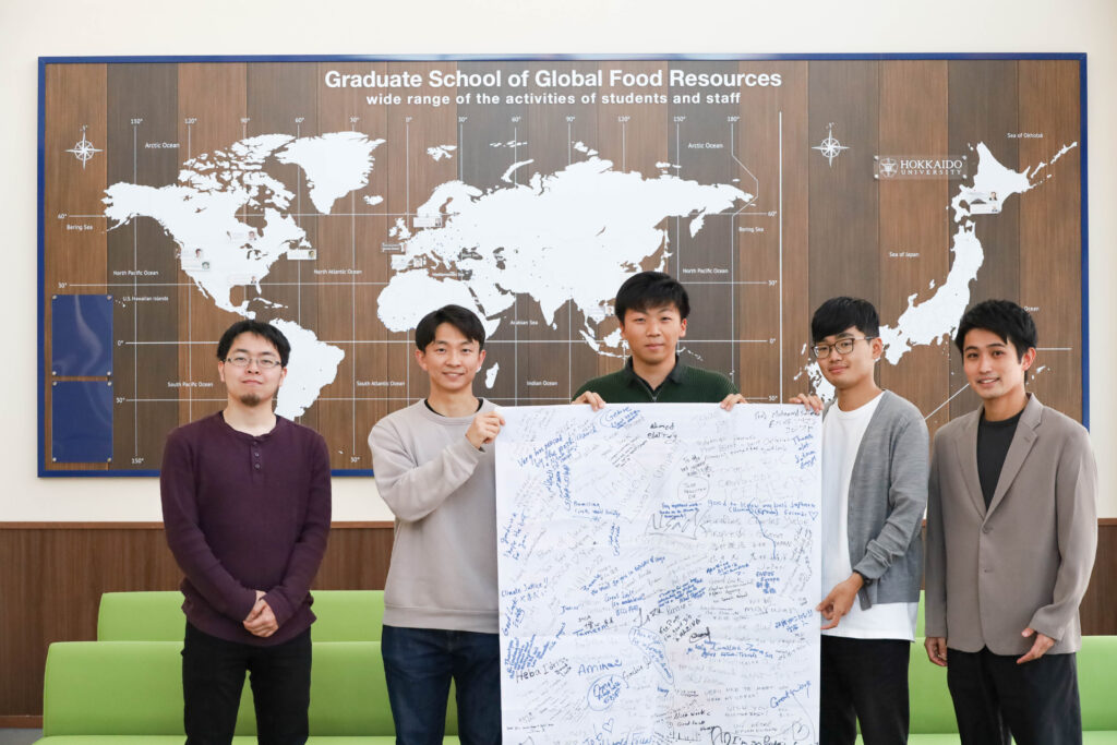 Five students are standing in a row in front of a world map facing to the camera for the picture. The three students in the middle are holding a big poster, showing it to the camera.