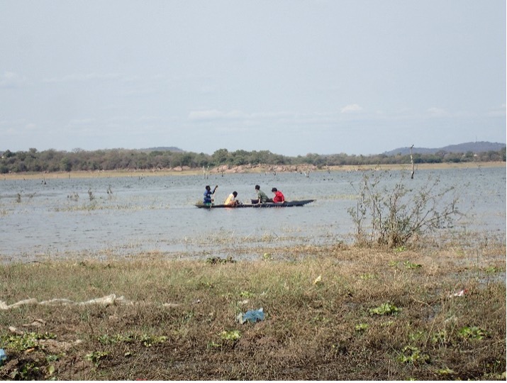 A photo taken from the land showing a lake. In the middle of the lake there is a boat carrying four people. Person on the far left is holding an oar.