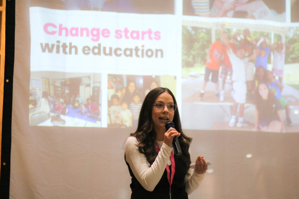 Carla Osnaya is holding a microphone, giving a presentation in front of a slideshow. The slideshow is showing "Change starts with education"