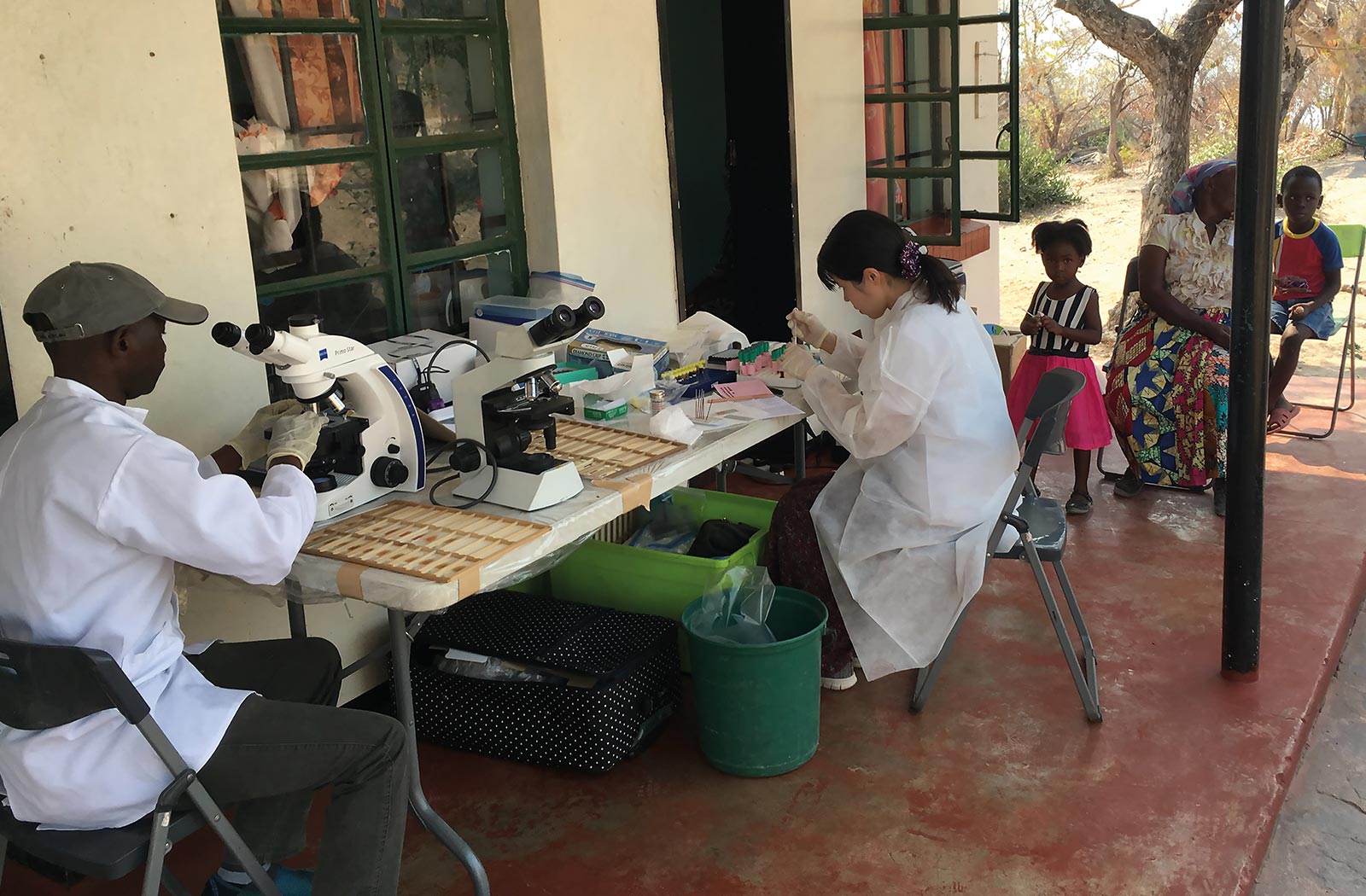 A photo of scientists performing microscopic observations for disease diagnosis in the outdoors. Photo taken in Zambia; provided by Kyoko Hayashida.