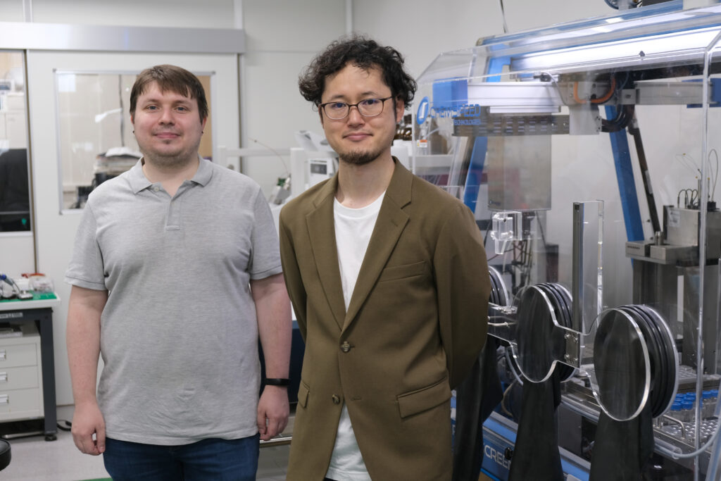 Pavel Sidorov (left) and Nobuya Tsuji (right) of the research team at the Institute for Chemical Reaction Design and Discovery (WPI-ICReDD), Hokkaido University, pictured next to the automated synthesis robot used in this study. (Photo Credit: ICReDD)
