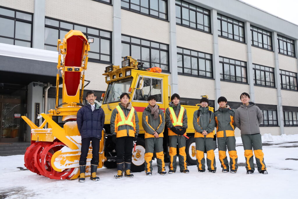 Associate Professor Takanori Emaru and his six students are standing in line posing for a picture in front of the snowplow used for a demonstration test. The campus' building is seen in the background.