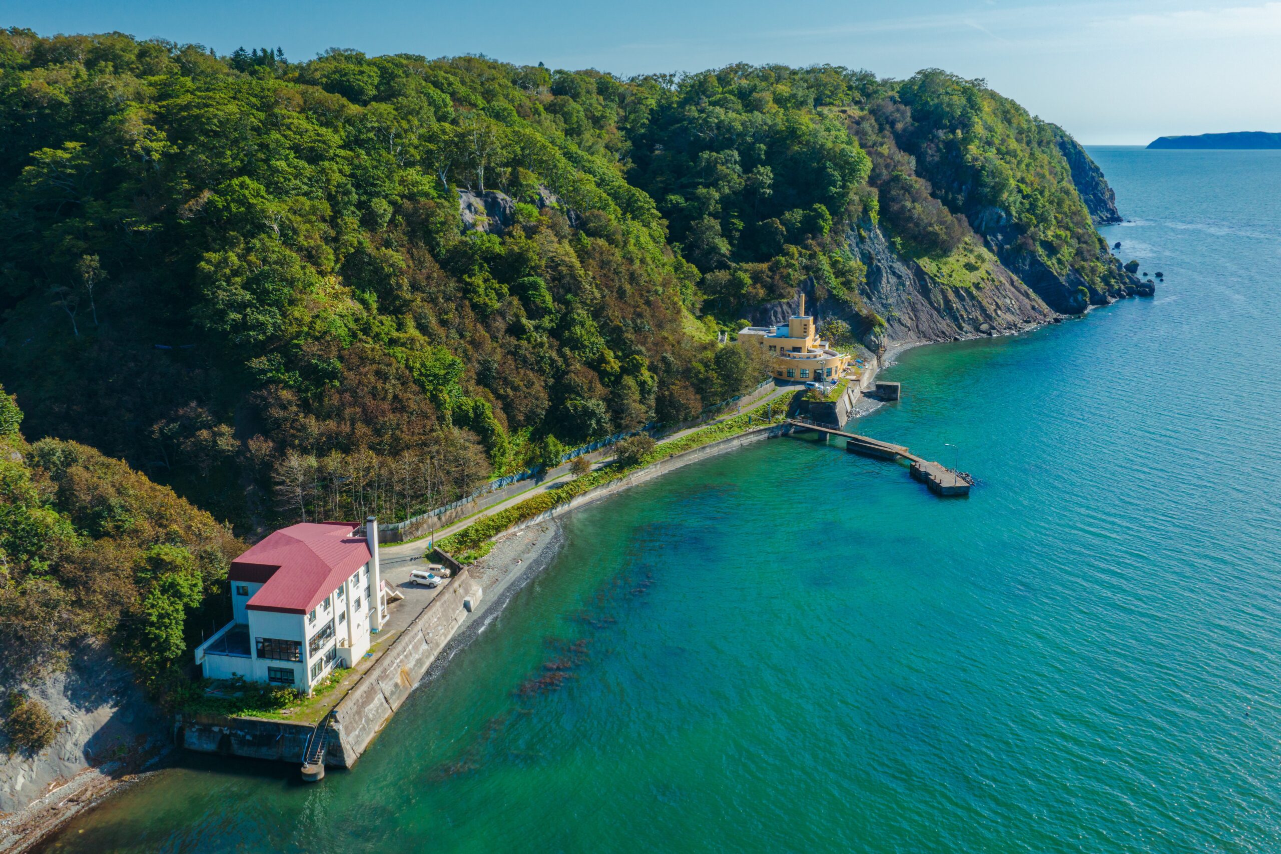 An aerial view of Hokkaido University’s Akkeshi Marine Station under the cliffs covered with greens. A white building is at the foreground and a yellow building is at the background, both connected by a road. A pier is in between. The land is facing the blue sea.