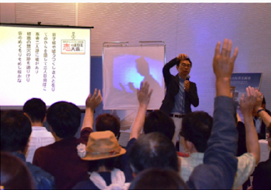 The scenery of voting for the best Haiku. A person standing in front of a large audience holding a microphone. Some people from the audience member are raising their hands.