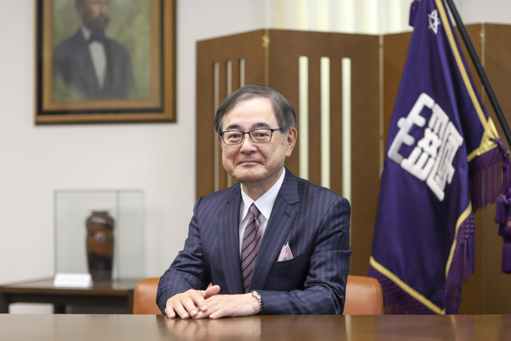 President Kiyohiro Houkin seated behind a desk. A purple university banner is behind him.