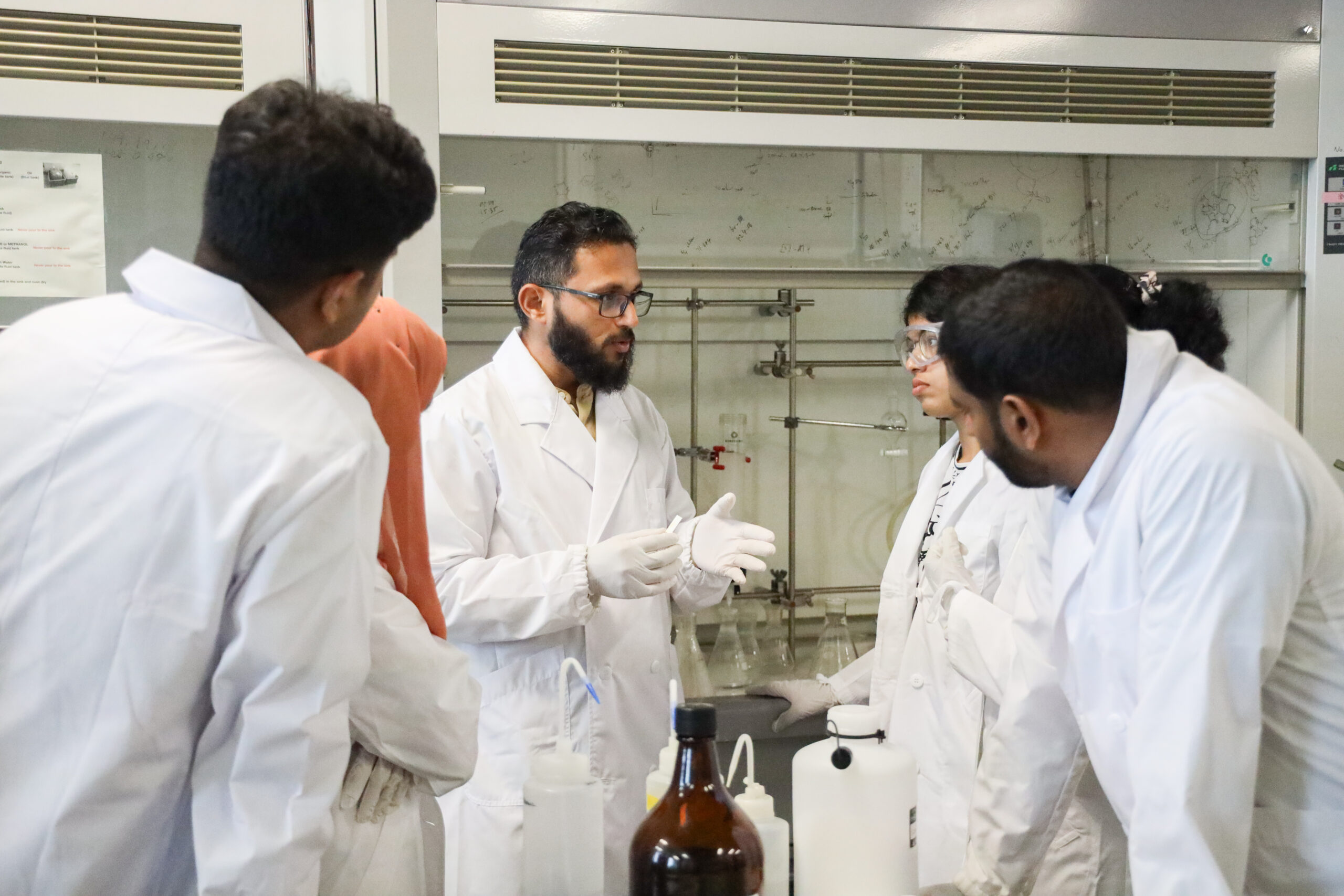 Dr. Hashim and 5 students in lab coats are in the middle of a discussion in a laboratory.