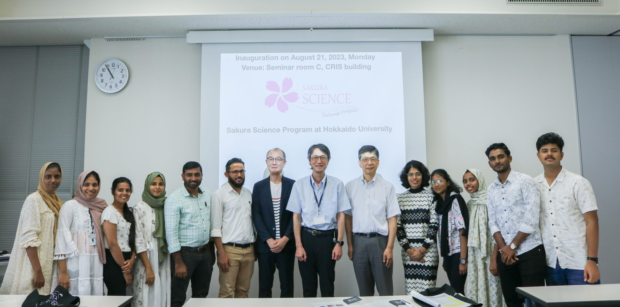Participants and the lecturers of Sakura Science at are standing in a single line, posing for the picture. A projected image of Sakura Science Exchange Program's logo can be seen on the background.