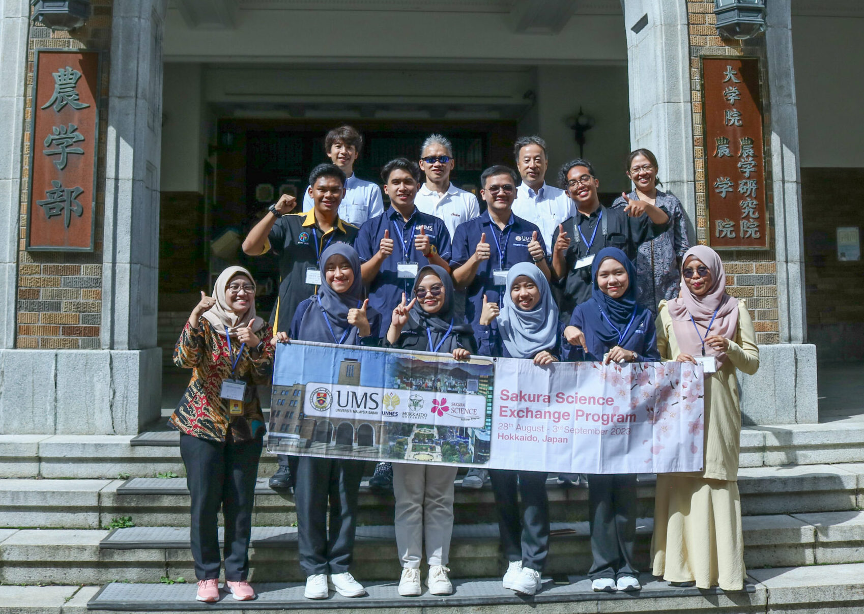 Participants and lecturers for the Sakura Science at the School of Agriculture are standing in front of the School of Agriculture's building's entrance, posing for the picture. The people on the front row are holding a banner of the program.