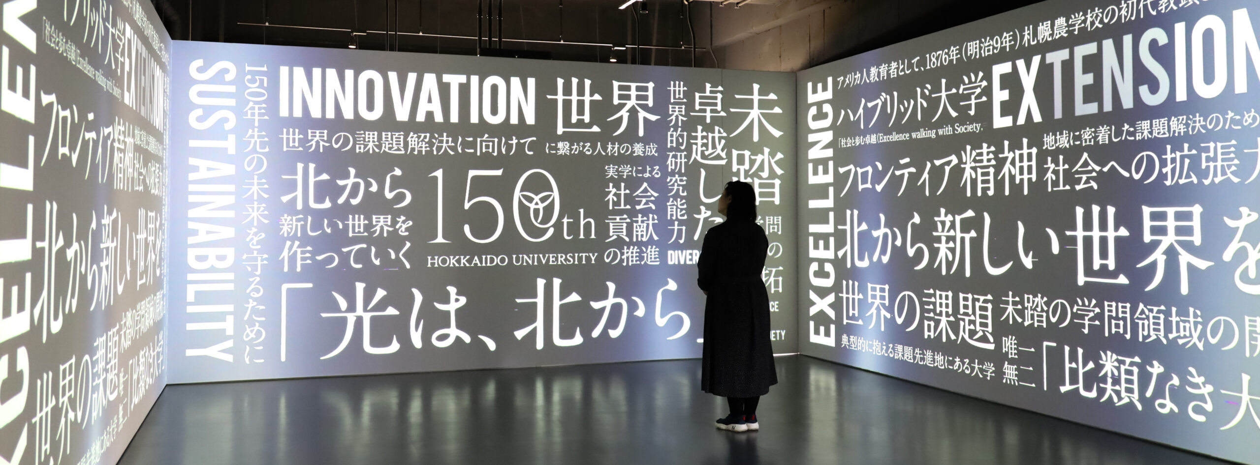 A person is standing in the middle of a three-wall projection room. The projection is displaying keywords in both English and Japanese on Hokkaido University's actions and visions.