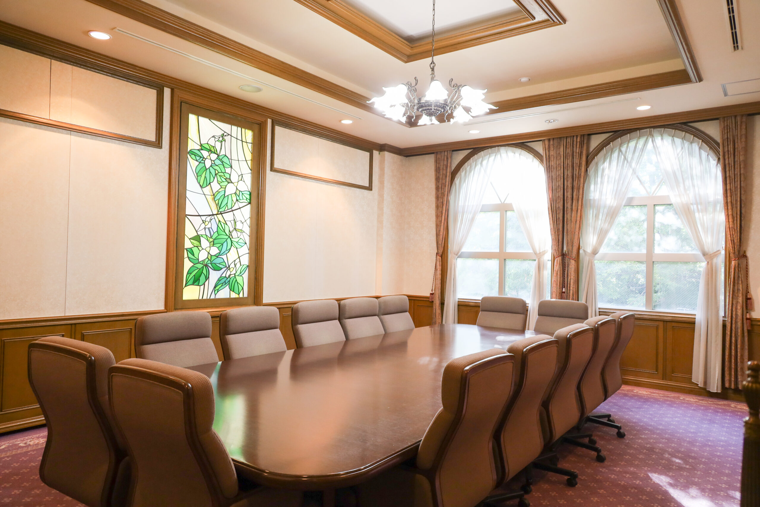 An interior look of the antique conference room. A rounded-rectangle-shaped conference table is sitting in the middle surrounded by conference chairs. On the left-side wall there is  a stained glass window portraying Trilliums.
